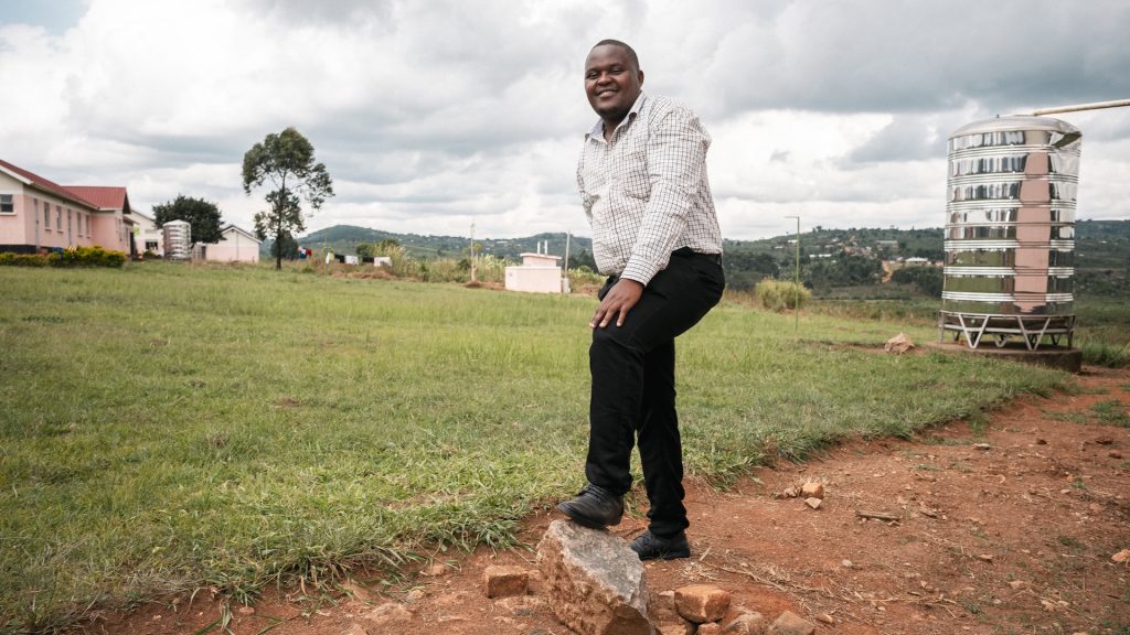 A man standing with one foot on a rock. Behind him is a silver water tank, a tree and a building.