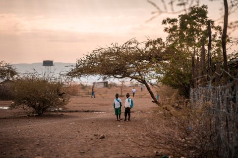 Kaksi koulupukuista lasta seisoo puun varjossa kenialaisella pakolaisalueella. Two children in school uniforms stand in the shade of a tree in a Kenyan refugee camp.