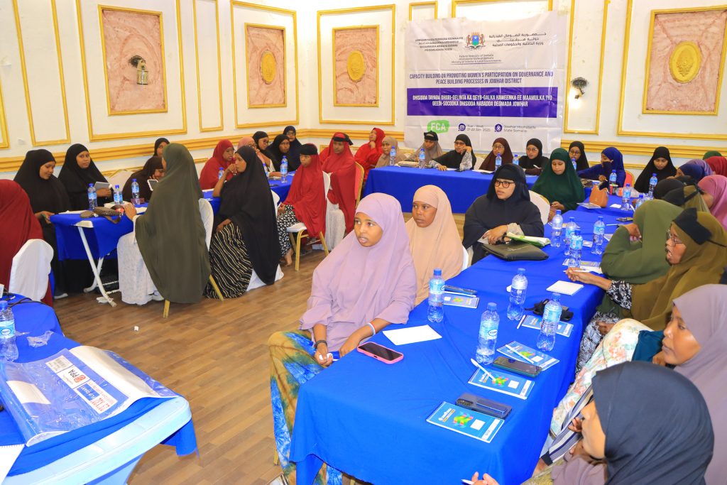 A large number of women sit at tables with notebooks, pens and water bootles in a meeting room. They are listening to someone off-camera speaking. On the wall is a banner with the logos of FCA, the Somali Ministry of Interior and the Finnish Ministry of Foreign Affairs.