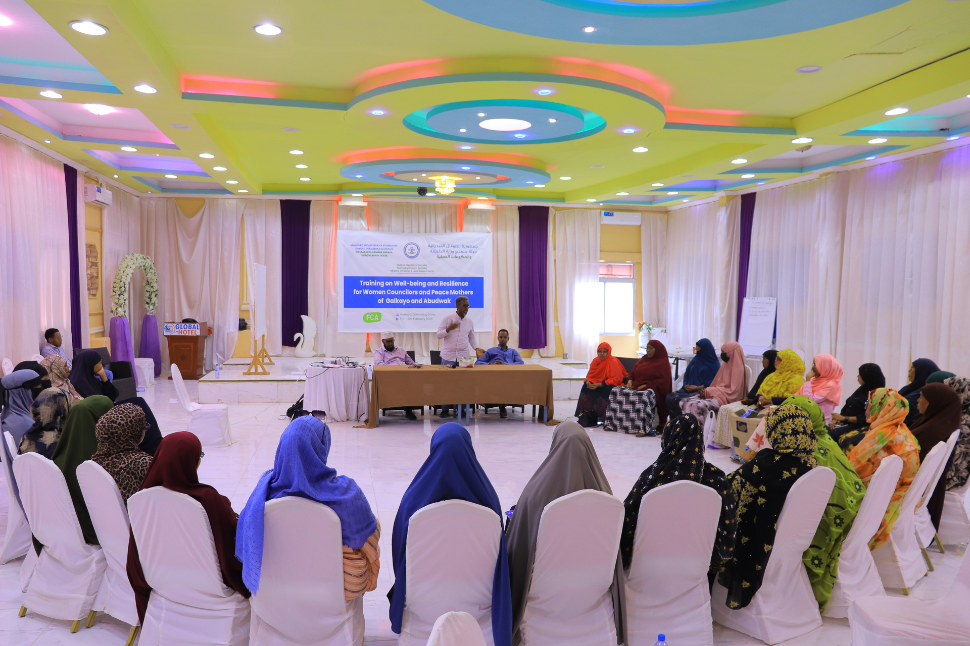 A group of women in a conference room sit in chairs arranged in a circle listening to a man at a desk talking.