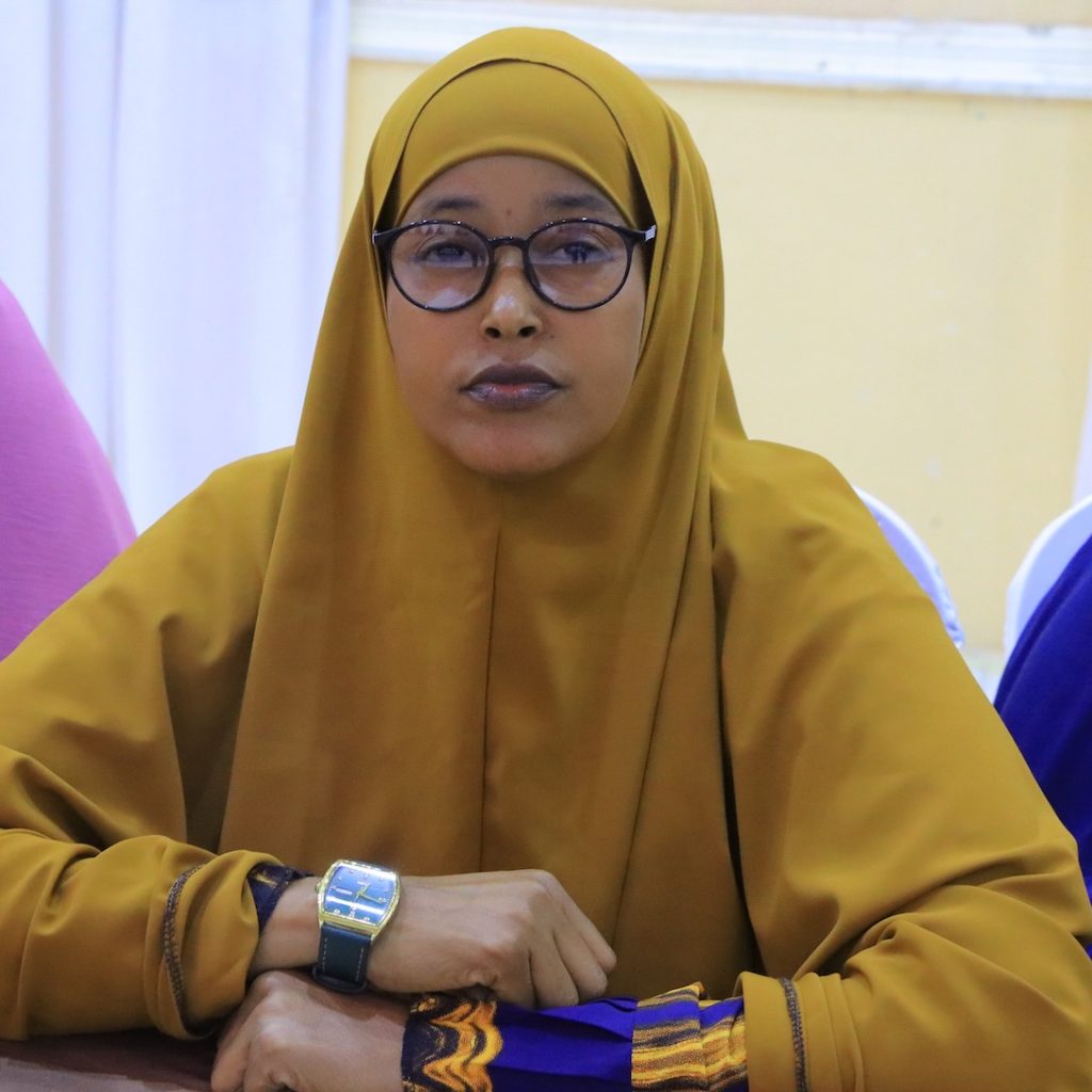 A woman in a yellow hijab sits at a table and looks directly into the camera.