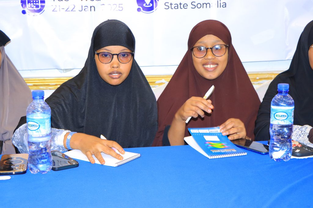 Two women sit at a table. They are holding pens and notebooks.