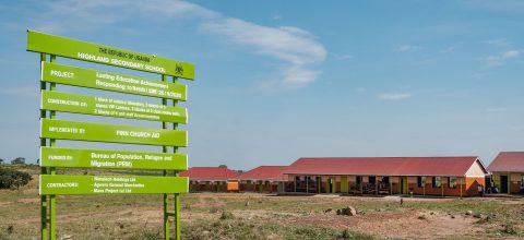 A sign in an open field in front of some school buildings.